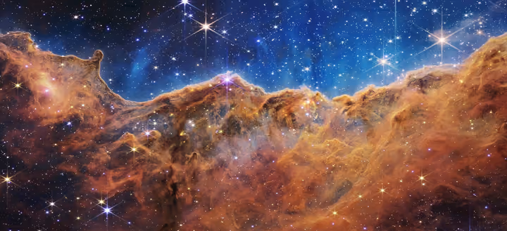 Ridge of glowing orange gas and dust in the Carina Nebula under a blue sky full of stars